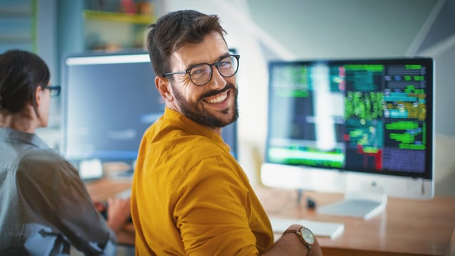 Smiling IT expert in front of monitor with IT monitoring dashboard - symbolic image for professional support and expert knowledge in IT monitoring with USU.