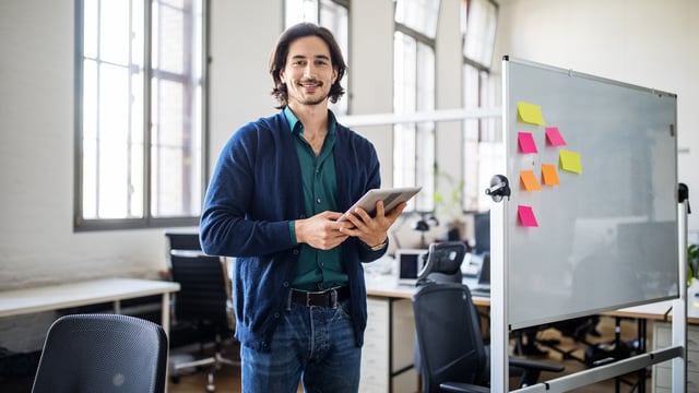 A USU IT Monitoring trainer stands in a modern office, holding a tablet and smiling confidently. A whiteboard with colorful sticky notes behind him suggests an active training or planning session focused on IT monitoring solutions.