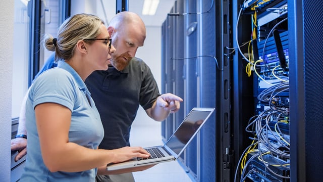 Two IT professionals working together in a server room. A woman is holding a laptop while a man points at the screen. Server racks with network cables and hardware are visible in the background.