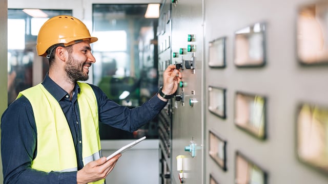 Technician with hard hat and tablet monitors switchgear in the control room. Symbolizes data center &amp; building infrastructure monitoring.