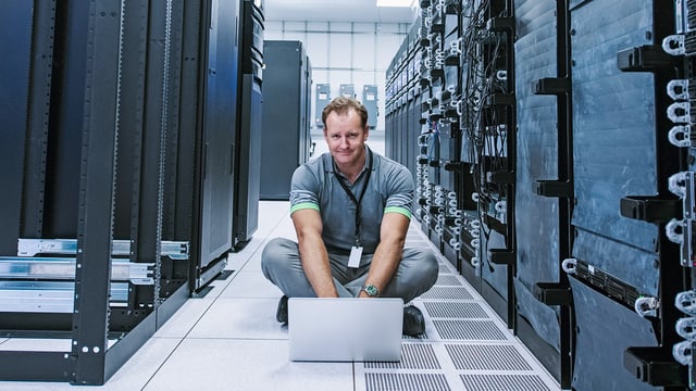 IT specialist sits with laptop in the server room in front of large server racks. Symbolizes mainframe monitoring.