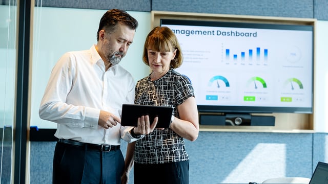 Two managers analyze capacity data on a tablet while a management dashboard is displayed in the background. Symbolizes capacity management