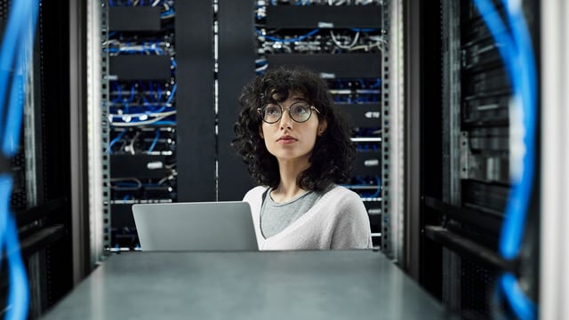 IT administrator with laptop in the server room, monitoring servers and network cables. Symbolizes USU Application & Server Monitoring (APM) for reliable IT monitoring. IT administrator with laptop in the server room, monitoring servers and network cables. Symbolizes USU Application & Server Monitoring (APM) for reliable IT monitoring.