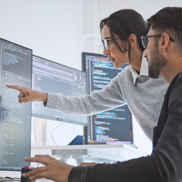 Two IT specialists analyze source code together on several monitors in order to actively manage IT risks.
